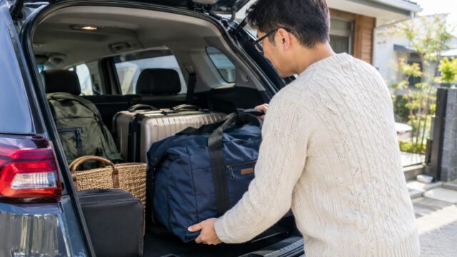 アイボリーのニットを着た眼鏡の父親が車のトランクに旅行バッグを積み込んでいる